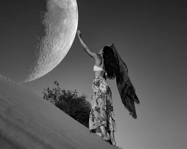 A woman with long hair reaches toward the large moon in the sky, standing on a slope with a shawl flowing behind her.