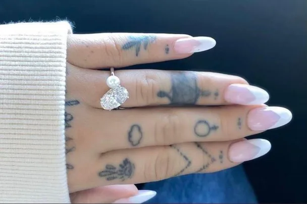 A hand with tattoos, wearing a ring with diamonds and pearls, and nails with a pink and white French manicure.