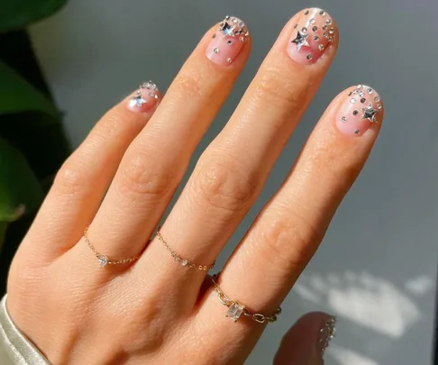 Close-up of a hand with nude nails adorned with silver star and dot embellishments suitable for New Year's Eve.