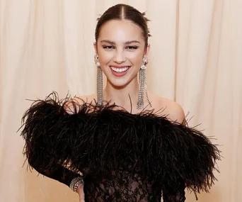 Smiling woman with sleek hair, wearing statement earrings and a black feathered outfit against a light background.