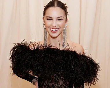 Smiling woman with sleek hair, wearing statement earrings and a black feathered outfit against a light background.