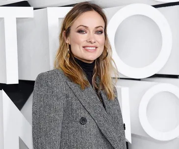 A woman in a herringbone coat smiles, standing in front of a geometric, monochrome background.