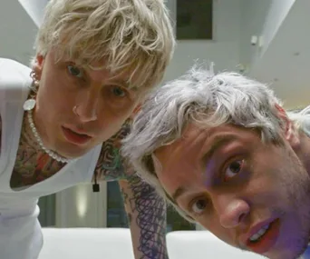 Two men with bleached hair look down at the camera indoors, one wearing a white tank top and jewelry.