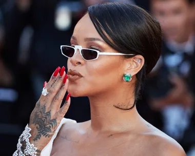 Person with short hair, sunglasses, red nails, and tattoos, blowing a kiss at an event, wearing a jeweled bracelet and earrings.