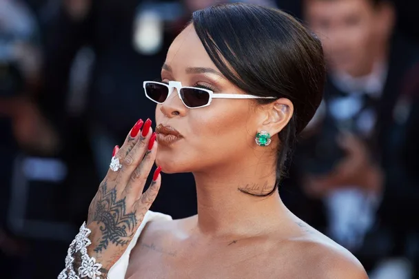 Person with short hair, sunglasses, red nails, and tattoos, blowing a kiss at an event, wearing a jeweled bracelet and earrings.