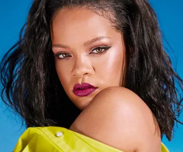 A woman with dark hair and vibrant makeup poses against a blue background wearing a bright yellow outfit.