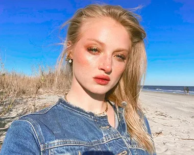 A person in a denim jacket poses on a sunny beach with grass and the ocean in the background.