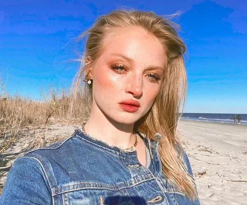 A person in a denim jacket poses on a sunny beach with grass and the ocean in the background.