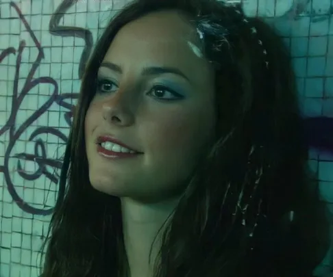 Young woman with long hair and makeup smiles against a graffiti-covered wall.