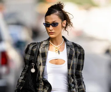 Woman in a checkered blazer and sunglasses walks on a city street carrying a black quilted handbag.