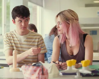 Two people sitting at a table with a model, one handing a condom to the other in a classroom setting.