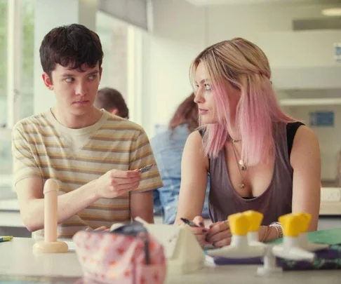 Two people sitting at a table with a model, one handing a condom to the other in a classroom setting.