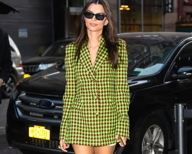 A person in a green and purple checkered blazer dress and sunglasses walks on a street near parked cars.