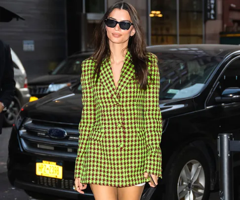 A person in a green and purple checkered blazer dress and sunglasses walks on a street near parked cars.