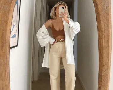 Woman taking a mirror selfie wearing an oversized white shirt, beige pants, and a brown top in a hallway.