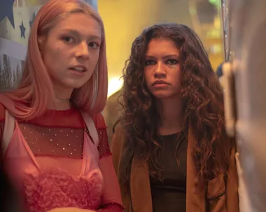 Two young individuals, one with pink hair and one with curly hair, stand closely in a dimly lit setting.