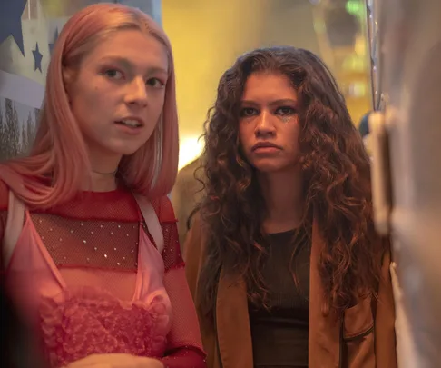 Two young individuals, one with pink hair and one with curly hair, stand closely in a dimly lit setting.