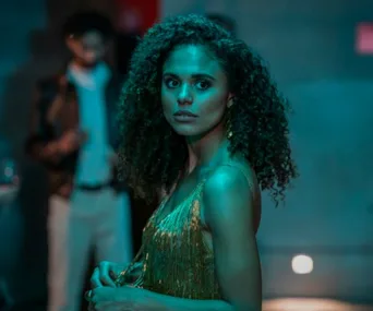 A woman with curly hair in a shimmering dress glances sideways in a dimly lit setting.