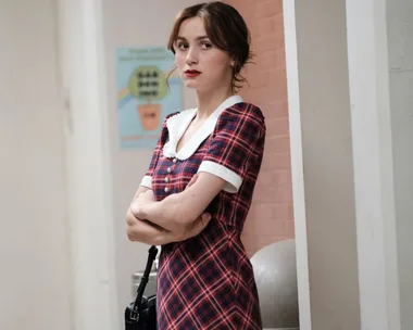 Person in a plaid dress with a white collar stands with arms crossed near a brick wall.