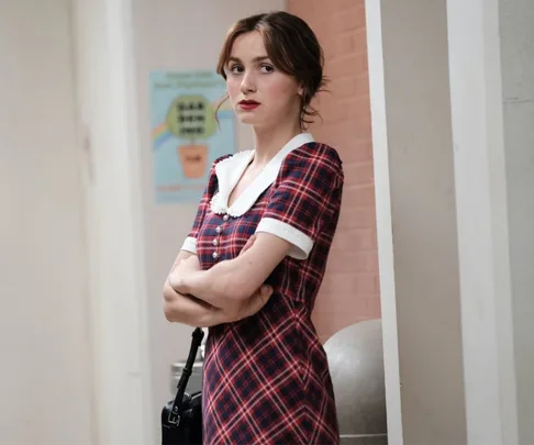 Person in a plaid dress with a white collar stands with arms crossed near a brick wall.