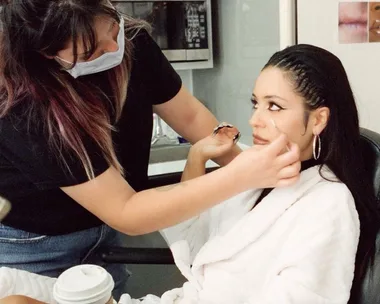 Makeup artist applies winged eyeliner to a woman in a white robe on set.