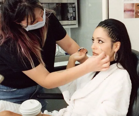 Makeup artist applies winged eyeliner to a woman in a white robe on set.