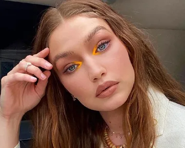 Woman with long hair and bold yellow eye makeup poses in a thoughtful manner, wearing natural lipstick.