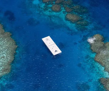 A floating tennis court with Adidas branding on crystal-clear blue ocean surrounded by coral reefs.