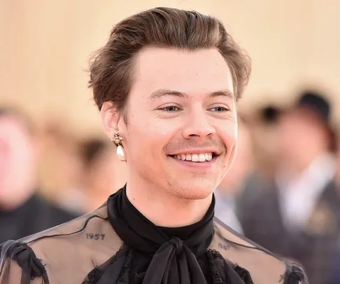 A smiling person with short hair and a pearl earring, wearing a semi-transparent black outfit with ruffled detail.