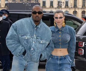 Two people in matching blue denim outfits on a street with a black vehicle and buildings in the background.