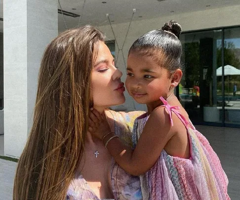 A woman with long hair kisses a young girl in a colorful dress outdoors.