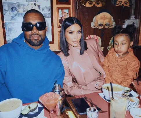 A family sitting at a restaurant table with food and drinks, featuring a man in sunglasses, a woman, and a child.