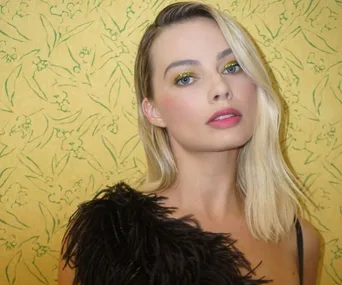 Blonde woman with green eye makeup, wearing a black feathered top, against a yellow floral background.