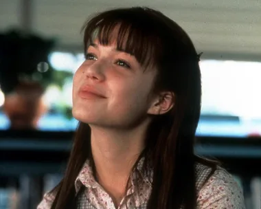 A young woman with long hair smiling softly, wearing a floral shirt, looks upward in a serene indoor setting.