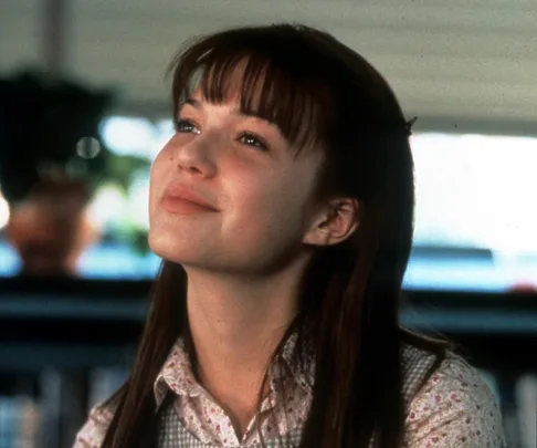 A young woman with long hair smiling softly, wearing a floral shirt, looks upward in a serene indoor setting.