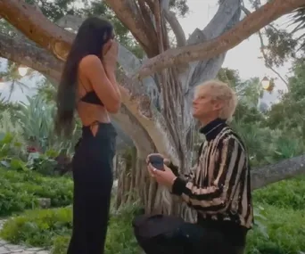 A person kneels, proposing to a surprised woman by a large tree in a garden setting.