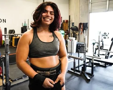 Person smiling in a gym, wearing a sports bra and preparing to lift weights.