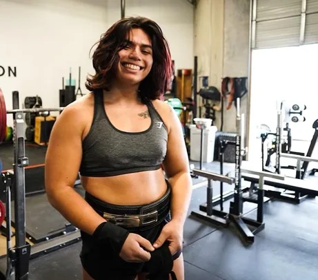 Person smiling in a gym, wearing a sports bra and preparing to lift weights.