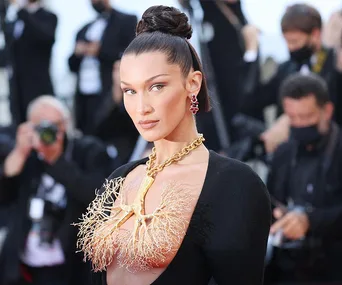 Bella Hadid at an event in a black dress with a gold lung-shaped neckline piece, surrounded by photographers.