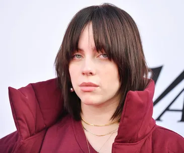 A person with medium-length dark hair, wearing a burgundy coat and gold necklaces, against a plain background.