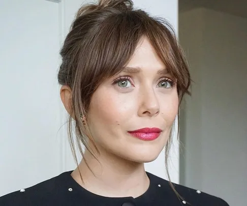 Woman with light brown bottleneck bangs, hair tied in a bun, wearing a black top and red lipstick, looking confidently.