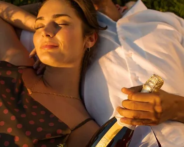 A woman relaxes on a sunny day with her head on a partner’s shoulder, holding a bottle of sparkling wine.