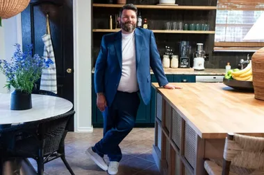 Man in a blue suit leaning against a kitchen counter, with a table and flowers on the left and kitchen items behind.