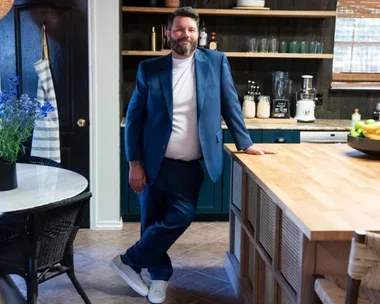 Man in a blue suit leaning against a kitchen counter, with a table and flowers on the left and kitchen items behind.