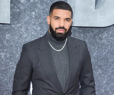 Man in a charcoal suit and turtleneck, wearing a necklace, stands against a textured gray background.