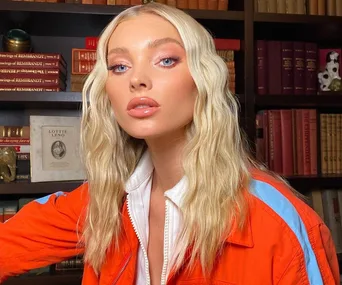 Blonde woman with long, wavy hair in an orange jacket in front of bookshelves.