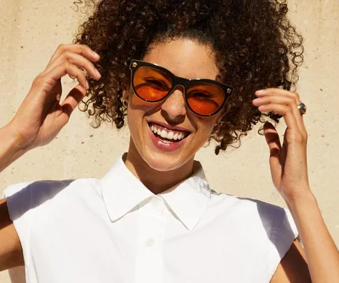 Woman with curly hair wearing orange-tinted sunglasses, white shirt, smiling against a beige background.