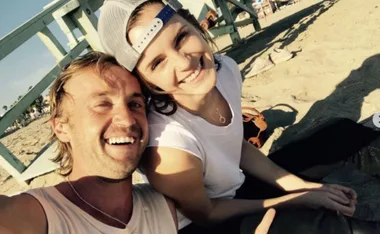 Two people smiling together on a sandy beach, one wearing a cap backward.