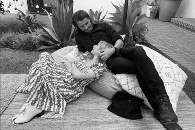 A couple relaxes on the patio, woman in floral dress rests her head on man's lap, surrounded by plants.