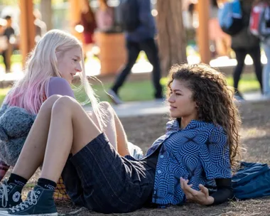 Two young women sit on the ground in a park, talking and laughing, with a blurred crowd in the background.